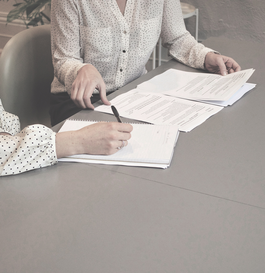 Two people studying a stack of documents laid out on a table and taking notes on a notebook